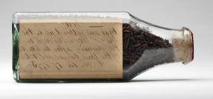 shriveled brown tea leaves sit inside a glass bottle 这是 closed up with a cork; cursive h和writing in brown ink on a yellowed paper is inside the bottle